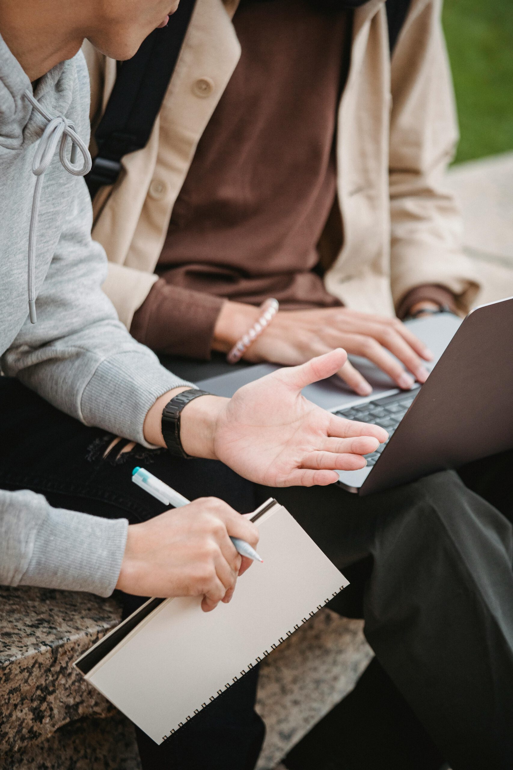 Two young adults collaborating with a laptop and notebook outdoors, discussing a project.